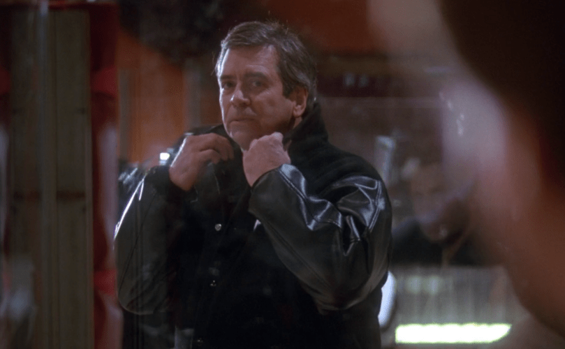 A man in a coat popping his collar with a smug look on his face.