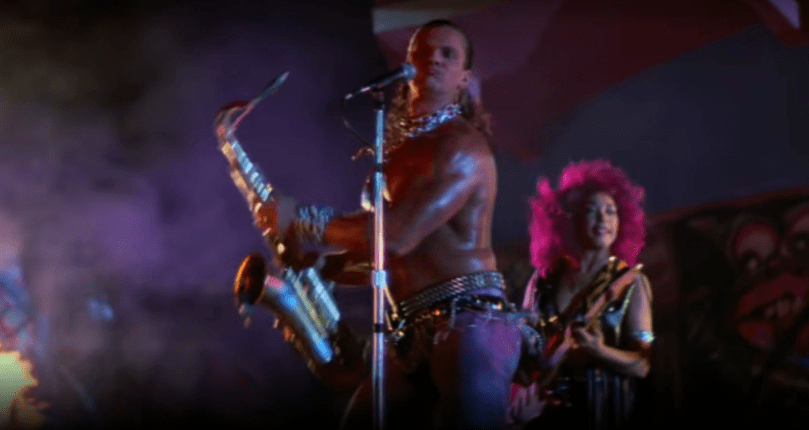Man dancing on stage with a saxophone while guitarist plays in the background